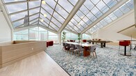 Located in the heart of downtown Cincinnati at 19 Seventh St W, this remarkable single-floor amenity space is defined by its breathtaking architectural centerpiece: a soaring, multi-panel glass skylight roof supported by sleek white structural beams that flood the interior with abundant natural light. Floor-to-ceiling glass walls frame sweeping panoramic views of the Cincinnati skyline, creating an airy, light-filled atmosphere throughout.

The expansive open layout is thoughtfully organized into distinct functional zones. A glazed conservatory-style area serves as the social hub, featuring a long communal dining table with mid-century modern chairs, high-top bar seating, and potted trees that add a refreshing natural element. Vibrant orange velvet accent chairs and a curved caramel leather sofa provide inviting lounge seating, complemented by a bold blue decorative area rug that anchors the space. A billiards table offers a recreational focal point, while a sculptural cylindrical architectural column adds a striking design statement. Built-in cabinetry with navy blue accents and light hardwood flooring complete the refined interior palette. The combination of natural light, elevated city views, and thoughtfully curated furnishings creates a sophisticated and versatile environment ideal for both social gatherings and relaxed everyday use.