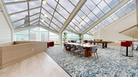 Located in the heart of downtown Cincinnati at 19 Seventh St W, this remarkable single-floor amenity space is defined by its breathtaking architectural centerpiece: a soaring, multi-panel glass skylight roof supported by sleek white structural beams that flood the interior with abundant natural light. Floor-to-ceiling glass walls frame sweeping panoramic views of the Cincinnati skyline, creating an airy, light-filled atmosphere throughout.

The expansive open layout is thoughtfully organized into distinct functional zones. A glazed conservatory-style area serves as the social hub, featuring a long communal dining table with mid-century modern chairs, high-top bar seating, and potted trees that add a refreshing natural element. Vibrant orange velvet accent chairs and a curved caramel leather sofa provide inviting lounge seating, complemented by a bold blue decorative area rug that anchors the space. A billiards table offers a recreational focal point, while a sculptural cylindrical architectural column adds a striking design statement. Built-in cabinetry with navy blue accents and light hardwood flooring complete the refined interior palette. The combination of natural light, elevated city views, and thoughtfully curated furnishings creates a sophisticated and versatile environment ideal for both social gatherings and relaxed everyday use.