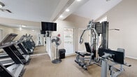Located on Sir Winston Street in San Antonio, TX, this community fitness center offers a well-equipped workout space designed to meet a variety of exercise needs. The room features vaulted, beamed ceilings that lend a sense of openness, with recessed lighting supplementing the natural light that streams in through several large windows fitted with blinds. Views of an outdoor courtyard with potted plants and a pool area can be glimpsed through the windows, creating a pleasant backdrop for workouts.

The fitness center is outfitted with a full multi-station cable weight machine as its centerpiece, enabling a wide range of strength training exercises. A dedicated area near the entrance houses two elliptical cross-trainers positioned side by side, while additional cardio equipment, including a treadmill, is visible throughout the space. A wall-mounted flat-screen television provides entertainment during workouts, and strategically placed mirrors amplify the sense of space and allow users to monitor their form.

The flooring is carpeted throughout, and safety essentials such as a fire extinguisher are present. The layout is thoughtfully organized, with clear pathways between equipment. A standing fan adds to the overall comfort of the environment. This fitness center serves as a convenient and functional amenity for residents in San Antonio.