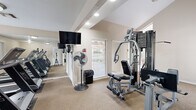 Located on Sir Winston Street in San Antonio, TX, this community fitness center offers a well-equipped workout space designed to meet a variety of exercise needs. The room features vaulted, beamed ceilings that lend a sense of openness, with recessed lighting supplementing the natural light that streams in through several large windows fitted with blinds. Views of an outdoor courtyard with potted plants and a pool area can be glimpsed through the windows, creating a pleasant backdrop for workouts.

The fitness center is outfitted with a full multi-station cable weight machine as its centerpiece, enabling a wide range of strength training exercises. A dedicated area near the entrance houses two elliptical cross-trainers positioned side by side, while additional cardio equipment, including a treadmill, is visible throughout the space. A wall-mounted flat-screen television provides entertainment during workouts, and strategically placed mirrors amplify the sense of space and allow users to monitor their form.

The flooring is carpeted throughout, and safety essentials such as a fire extinguisher are present. The layout is thoughtfully organized, with clear pathways between equipment. A standing fan adds to the overall comfort of the environment. This fitness center serves as a convenient and functional amenity for residents in San Antonio.