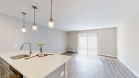 This efficient single-floor unit offers a thoughtfully designed layout that maximizes its compact footprint. The combined kitchen and living area forms the heart of the space, where a sleek, light-toned quartz peninsula takes center stage, illuminated by a trio of elegant glass pendant lights. The kitchen is equipped with stainless steel appliances, including a refrigerator and microwave, complemented by gray cabinetry and recessed lighting. The open living area flows seamlessly from the kitchen, featuring light hardwood-style flooring and neutral, warm-toned walls that create a bright and airy ambiance. A large sliding glass door dressed with vertical blinds allows abundant natural light to pour in, adding to the open feel of the space. Two bathrooms serve the unit, each featuring wood-style flooring and clean, neutral finishes. One bathroom includes a glass-enclosed walk-in shower with classic white subway tile, providing a sleek, modern aesthetic. Two hallways connect the various areas of the unit, ensuring an efficient flow throughout. The consistent use of light flooring, neutral wall tones, and modern fixtures throughout the space creates a cohesive, contemporary look that is both functional and inviting.