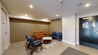 Located at 62 Armstrong Court in Halifax, Nova Scotia, this building lobby offers a polished and well-appointed entry experience. The space is thoughtfully designed to balance functionality with aesthetic appeal, making a strong first impression for residents and visitors alike.

The focal point of the lobby is a contemporary seating area anchored by a geometric-patterned area rug over light tile flooring. Mid-century modern furnishings — including a cognac leather loveseat and a set of navy blue armchairs with walnut-toned frames — are arranged around a marble-topped oval coffee table, creating an inviting lounge atmosphere. A full-height slatted wood accent wall runs along one side, adding warmth and texture to the otherwise clean, white interior.

Recessed ceiling lighting provides consistent, ambient illumination throughout. The elevator bank is a standout design feature, with two stainless steel elevator doors framed by bold dark chevron-patterned tile, lending a refined, contemporary edge to the space.

The lobby benefits from natural light filtering in through the glass entry doors, which open directly to the street. Durable, low-pile carpet defines the main entry corridor, transitioning to the tiled lounge zone. The overall design is clean, modern, and welcoming — reflecting a well-maintained residential building in Halifax.