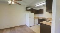 This single-floor space features an open-concept layout that connects the kitchen and living area seamlessly. The kitchen is equipped with dark brown cabinetry mounted along the upper walls, providing ample storage. A light-toned laminate countertop runs along the base cabinets, offering functional workspace. White appliances, including a full-size refrigerator, dishwasher, and a stovetop, are neatly integrated into the kitchen layout. A double sink is positioned along one wall, adding practicality to the space. The flooring in the kitchen area transitions from light vinyl tile to carpeting in the adjacent living space, clearly delineating the two zones. The living area is modestly sized and benefits from a ceiling fan, which aids in air circulation. Overhead fluorescent lighting in the kitchen ensures well-lit meal preparation. The textured ceiling adds a classic touch throughout the space. The overall design is straightforward and functional, with neutral wall tones that offer a clean, understated backdrop suitable for personalizing with furnishings and décor.