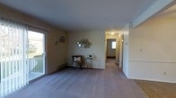 This single-floor residence offers a straightforward and functional layout designed for comfortable everyday living. The main living area is finished with wall-to-wall carpeting in a neutral tone, contributing to a warm and quiet atmosphere throughout. A large sliding glass door fills the space with natural light and provides direct access to an outdoor balcony or patio area, offering pleasant exterior views. A wall-mounted air conditioning unit ensures year-round climate comfort, while a baseboard heating system runs along the perimeter walls for efficient warmth during cooler months. Decorative touches, including a mirrored wall accent and a patterned accent chair with a coordinating side table, add character to the living space. A hallway extends from the main living area, leading to what appears to be additional rooms including bedrooms and a bathroom, each accessible via individual doors. The corridor is well-lit with overhead lighting and features a window at the far end, drawing additional natural light into the interior. The overall color palette is soft and neutral, with light-toned walls and ceilings that enhance the sense of openness. The layout is practical and cohesive, offering a comfortable single-level living experience.