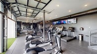 Located in Columbus, OH, this well-appointed fitness center offers a comprehensive workout environment designed with both functionality and aesthetics in mind. The space is finished in a contemporary dark-and-grey palette, with charcoal walls, exposed ductwork overhead, and durable rubber-tile flooring throughout, creating an industrial yet polished atmosphere.

The cardio zone is prominently featured along one wall, lined with a full row of treadmills, ellipticals, and stair climbers from Life Fitness, all positioned in front of a bank of wall-mounted flat-screen TVs for an engaging workout experience. Adjacent to the cardio area, a dedicated cycling and rowing section provides additional low-impact training options, benefiting from abundant natural light streaming through multiple windows along the exterior wall.

A separate strength training area houses a well-stocked dumbbell rack with a complete range of weights, kettlebells, a weight bench, and a cable machine, giving users everything needed for a full resistance training session. Full-length mirrors amplify the sense of space and allow for proper form checks throughout.

One of the most distinctive architectural features is the large glass-panel garage-style door that opens to a private outdoor turf area, seamlessly blending indoor and outdoor fitness. Recessed lighting and steel structural columns complete the modern industrial design of this fully equipped facility.