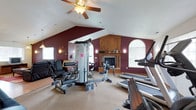 This dedicated fitness space is designed to deliver a full workout experience within a thoughtfully appointed interior. The room immediately draws attention with its vaulted, cathedral-style ceiling, which lends an open and airy feel, complemented by a ceiling fan that provides comfortable airflow during workouts. Bold burgundy accent walls provide a striking contrast against the crisp white ceiling and trim, creating an energizing yet refined ambiance.

A wood-burning fireplace, framed by a handsome wooden mantel with arched detailing, serves as the room's architectural focal point, adding warmth and character typically not found in a standard fitness environment. Flanking the fireplace, arched windows allow natural light to fill the space, enhancing the overall atmosphere.

The fitness area is well-stocked with equipment, including a multi-station cable strength machine, a stationary bike, and a treadmill, offering a comprehensive range of cardiovascular and resistance training options. Wall-mounted sconces add subtle accent lighting throughout. Neutral carpeting underfoot keeps the space comfortable, and the generous open floor plan allows for easy movement between equipment. This is a truly functional and inviting personal gym.