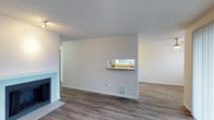 This single-floor residence offers a clean, functional layout designed for comfortable everyday living. The main living area is finished with durable hardwood-style flooring in a warm wood tone that extends throughout the space, lending a cohesive and inviting feel. A classic fireplace anchors one side of the living room, serving as a natural focal point and adding a cozy character to the area.

A defining architectural feature is the pass-through window connecting the kitchen to the main living space. Fitted with a convenient countertop ledge, this opening allows natural light from the kitchen to filter into the living area while maintaining a sense of openness and connectivity between the two spaces. The kitchen, visible through the pass-through, includes standard appliances and a sink beneath a window that brings in additional natural light.

The walls are painted in a neutral off-white tone, complementing the light-toned baseboards and trim throughout. A textured popcorn ceiling adds a classic residential finish overhead. A floor-level wall vent indicates a central heating system integrated into the space. The overall ambiance is bright, neutral, and move-in ready, offering a versatile canvas for a variety of interior styles.