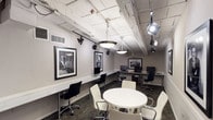 This well-appointed single-floor space functions as a professional business center and collaborative workspace, thoughtfully designed to blend functionality with visual character. The room features an extended marble-topped workstation along one wall, accommodating multiple desktop computers and ergonomic task chairs, making it well-suited for individual productivity. A central round white table surrounded by modern white upholstered chairs provides a comfortable setting for group meetings or collaborative work.

The industrial-inspired ceiling is a defining architectural element, showcasing exposed white-painted beams, ductwork, and visible piping overhead. Dramatic track lighting and large pendant fixtures cast a warm, focused glow throughout the space, adding both practicality and visual interest. The patterned carpet in deep teal and brown tones grounds the room and adds a layer of sophisticated texture.

Curated black-and-white photographic artwork is displayed prominently across the walls, lending the space a distinguished and cultured ambiance. Additional workstations along a secondary wall provide further computing resources. A richly stained wooden door with a small window offers access to an adjacent area. The neutral gray walls and white wainscoting tie together the overall design, creating a polished yet creative environment ideal for professional use.