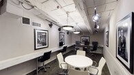 This well-appointed single-floor space functions as a professional business center and collaborative workspace, thoughtfully designed to blend functionality with visual character. The room features an extended marble-topped workstation along one wall, accommodating multiple desktop computers and ergonomic task chairs, making it well-suited for individual productivity. A central round white table surrounded by modern white upholstered chairs provides a comfortable setting for group meetings or collaborative work.

The industrial-inspired ceiling is a defining architectural element, showcasing exposed white-painted beams, ductwork, and visible piping overhead. Dramatic track lighting and large pendant fixtures cast a warm, focused glow throughout the space, adding both practicality and visual interest. The patterned carpet in deep teal and brown tones grounds the room and adds a layer of sophisticated texture.

Curated black-and-white photographic artwork is displayed prominently across the walls, lending the space a distinguished and cultured ambiance. Additional workstations along a secondary wall provide further computing resources. A richly stained wooden door with a small window offers access to an adjacent area. The neutral gray walls and white wainscoting tie together the overall design, creating a polished yet creative environment ideal for professional use.