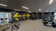 This fitness center is designed to support a full range of workout routines within a clean, contemporary space. The facility is organized into distinct functional zones, allowing users to move efficiently between different training styles. The main open area features carpeted flooring and is anchored by a striking dark gray accent wall adorned with bold yellow directional arrows, giving the space a dynamic, motivating aesthetic. Linear LED lighting runs across the ceiling throughout, providing consistent, bright illumination.

A dedicated cardio zone houses multiple treadmills, elliptical trainers, stationary bikes, and recumbent bikes, accompanied by a wall-mounted television for entertainment during workouts. Natural light filters in through windows along one wall, enhancing the overall atmosphere. Adjacent to this area, a functional training nook is stocked with medicine balls in various sizes, resistance bands, TRX suspension straps, foam rollers, and exercise balls displayed on a vertical rack.

The free weights section includes a dumbbell rack with an adjustable weight bench and a water station nearby for convenience. Posted fitness guidelines are visible on the wall, reflecting an organized and user-focused environment. Overall, the space balances energetic design elements with practical equipment placement.