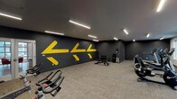This fitness center is designed to support a full range of workout routines within a clean, contemporary space. The facility is organized into distinct functional zones, allowing users to move efficiently between different training styles. The main open area features carpeted flooring and is anchored by a striking dark gray accent wall adorned with bold yellow directional arrows, giving the space a dynamic, motivating aesthetic. Linear LED lighting runs across the ceiling throughout, providing consistent, bright illumination.

A dedicated cardio zone houses multiple treadmills, elliptical trainers, stationary bikes, and recumbent bikes, accompanied by a wall-mounted television for entertainment during workouts. Natural light filters in through windows along one wall, enhancing the overall atmosphere. Adjacent to this area, a functional training nook is stocked with medicine balls in various sizes, resistance bands, TRX suspension straps, foam rollers, and exercise balls displayed on a vertical rack.

The free weights section includes a dumbbell rack with an adjustable weight bench and a water station nearby for convenience. Posted fitness guidelines are visible on the wall, reflecting an organized and user-focused environment. Overall, the space balances energetic design elements with practical equipment placement.