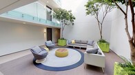 This common area is a thoughtfully designed indoor courtyard that combines architectural elegance with a welcoming, social atmosphere. The space rises to a double-height ceiling, with a glass-railed upper-level walkway that allows natural light to filter down and creates a sense of openness and vertical drama. Mature trees planted in vibrant lime-green planters anchor the space, introducing organic life and a refreshing natural element to the contemporary interior. The flooring transitions between neutral carpeting and hard surfaces, grounding the area with warmth and texture. At the center of the lounge zone, a large circular area rug with a navy border defines a comfortable gathering space furnished with a woven yellow ottoman-style coffee table featuring a wood-top surface. Seating includes a generously sized L-shaped gray sectional sofa adorned with patterned throw pillows, complemented by a pair of sculptural wicker swivel chairs with coordinating blue-and-white cushions. Wall sconces provide soft ambient lighting alongside recessed ceiling fixtures, contributing to a balanced and inviting glow. Unit entry doors with numbered signage are visible along the perimeter, integrating residential access seamlessly into this shared social hub.