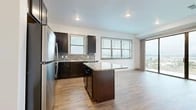 This well-designed single-floor unit offers a thoughtful layout with a clean, contemporary aesthetic throughout. The kitchen serves as a standout feature, showcasing dark espresso cabinetry, granite countertops, a textured tile backsplash, and a full suite of stainless steel appliances including a refrigerator, range, microwave, and dishwasher. A central kitchen island provides additional prep and seating space, while light wood-look flooring ties the kitchen seamlessly into the adjacent living area. The living space is bright and open, benefiting from multiple windows and access to a balcony that brings in natural light and outdoor views. A corridor with recessed lighting and wood-look flooring connects the main living areas to the private rooms. The bedrooms are finished with plush carpeting and feature windows that frame scenic water views. Paneled doors and crisp white walls maintain a consistent, modern feel throughout. The bathroom is well-appointed with a soaking tub, a separate glass-enclosed shower with white tile surround, wood-look flooring, and a dark vanity with granite countertops. A generously sized walk-in closet with built-in shelving and hanging rods provides ample storage, completing this functional and stylish unit.