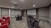 This elegantly designed media and lounge space combines cinematic functionality with upscale interior design. The room is anchored by a large projection screen paired with a ceiling-mounted projector, framed by floor-length drape panels that add a theatrical touch. Recessed lighting throughout illuminates the space evenly, complementing the refined wainscoting and paneled wall treatments finished in warm neutral tones with contrasting dark trim.

The flooring features a distinctive patterned carpet with an animal-print motif, lending a sophisticated visual texture underfoot. Seating is thoughtfully arranged to serve multiple purposes: bold red leather armchairs with chevron accent pillows provide vibrant focal points, while a wraparound tufted white banquette along one wall offers generous communal seating. Round pedestal tables in matte black are positioned throughout to accompany the various seating clusters.

A framed wall-mounted art installation of sculptural floral forms in gold tones serves as a standout decorative element above the banquette. A narrow sidelight window introduces a subtle touch of natural light along one wall. The overall ambiance is that of a private club or upscale entertainment lounge — stylish, comfortable, and well-appointed for both intimate gatherings and private screenings.