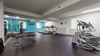 This fitness center offers a comprehensive workout environment designed to accommodate a variety of exercise routines. The space is laid out across a single floor and is defined by crisp white walls accented by bold teal feature walls, creating an energizing atmosphere. Durable rubber flooring covers the entire area, providing cushioning and safety for all types of workouts.

The cardio zone is equipped with multiple treadmills and a stationary bike, positioned near natural-light-filled windows that brighten the space. A stair climber and elliptical machines round out the cardio offerings. For strength training, the space includes a multi-function cable machine, a weight bench with a dumbbell rack, and an adjustable workout bench alongside a stability ball.

A large wall-mounted mirror spans one section of the room, enhancing the sense of space and allowing users to monitor their form during exercises. Recessed and overhead fluorescent lighting ensures the entire area is well-lit throughout the day. The layout includes additional open floor space ideal for stretching or bodyweight exercises. Two interior doors provide access to adjacent areas, and wall-mounted amenities such as a towel bar and a hand sanitizer dispenser add practical convenience to the facility.