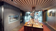 This business center is a thoughtfully designed single-level workspace that blends functionality with distinctive aesthetic character. The ceiling is a standout architectural feature, composed of alternating wood slats that add warmth and visual rhythm to the room. A long, solid wood communal work table runs the length of the space, outfitted with iMac computers and leather mouse pads, providing a productive environment for focused work or collaboration. Overhead, a series of industrial-style articulating pendant lamps are suspended from a central track, casting focused task lighting across the work surface. One wall is clad in dark tile and anchors a sleek, linear gas fireplace, adding an unexpected element of comfort and ambiance to the professional setting. Sheer floor-to-ceiling curtains frame large windows and a glass door along the exterior wall, flooding the space with natural light and offering views to an outdoor area beyond. The flooring features wood-tone planks with a vibrant area rug beneath the work table, grounding the space with color and texture. Decorative artwork, including a framed vintage typewriter print, adds a creative and curated touch that completes the refined yet inviting atmosphere of this well-appointed business center.