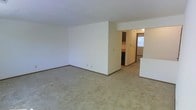 This single-floor residential unit offers a straightforward and functional layout designed to maximize usable living space. The main living area is generously proportioned, featuring light-toned carpet flooring and clean white walls that create a bright, neutral backdrop. A partial wall adds subtle architectural definition to the space, separating the living area from the rest of the unit while maintaining an open feel.

Toward the rear of the unit, a passageway leads into the kitchen, which is equipped with dark cabinetry and a window that invites natural light into the space. A glimpse of a door beyond suggests additional access points or adjoining rooms. To the side, a partial countertop or breakfast bar hints at a practical transition zone between the living and kitchen areas.

The overall ambiance is clean, simple, and move-in ready, with a neutral color palette throughout that lends itself well to a variety of décor styles. The layout flows in a logical and connected manner, making efficient use of the available space across the single level. The combination of carpeted common areas and the kitchen's functional design provides a comfortable and practical living environment.