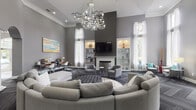 Located at 11010 Eastlake Club Dr in Raleigh, NC, this sophisticated single-floor clubhouse makes an immediate impression with its grand entrance — ornate leaded glass French doors topped by an arched transom window that floods the foyer with natural light. The interior is defined by soaring ceilings that lend an airy, expansive feel to the main lounge area, which is anchored by a striking sculptural chandelier and elegant white decorative columns framing graceful arched openings.

The primary gathering space is thoughtfully arranged around a curved sectional sofa and tufted ottoman, complemented by accent chairs in soft gray tones with teal accent pillows. A central fireplace with a classic white surround serves as a focal point, flanked by illuminated built-in shelving displaying curated décor. Large floor-to-ceiling windows dressed in sheer white curtains invite abundant natural light and offer views of the surrounding greenery.

The herringbone-patterned dark carpet throughout adds texture and sophistication to the neutral gray color palette. A dedicated workstation area with computer desks and task lighting provides a functional workspace, while colorful artwork adds vibrant contrast to the refined, contemporary interior. The overall ambiance is polished and welcoming, blending classic architectural details with modern furnishings.