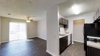 This single-floor residence offers a well-designed open-concept layout that seamlessly connects the kitchen and living areas, creating a functional and cohesive living space. Dark hardwood-style flooring runs throughout, providing a warm and grounded aesthetic that contrasts elegantly with the neutral white walls and textured ceilings.

The kitchen is positioned adjacent to the main living area, partially separated by a structural half-wall that defines the space without closing it off. Dark espresso-toned cabinetry lines the upper and lower sections of the kitchen, complemented by a light-colored countertop, a stainless steel dishwasher, and a built-in sink. A skylight-style ceiling fixture provides ample overhead illumination.

The living area is generously sized, featuring a ceiling fan with integrated lighting that adds both comfort and functionality. Vertical blinds cover a large sliding glass door or window along one wall, allowing for adjustable natural light and lending the room an airy, open feel. The textured ceiling adds a classic residential touch throughout the home. A doorway visible beyond the kitchen hints at additional rooms, rounding out this practical and inviting single-level layout.
