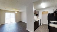 This single-floor residence offers a well-designed open-concept layout that seamlessly connects the kitchen and living areas, creating a functional and cohesive living space. Dark hardwood-style flooring runs throughout, providing a warm and grounded aesthetic that contrasts elegantly with the neutral white walls and textured ceilings.

The kitchen is positioned adjacent to the main living area, partially separated by a structural half-wall that defines the space without closing it off. Dark espresso-toned cabinetry lines the upper and lower sections of the kitchen, complemented by a light-colored countertop, a stainless steel dishwasher, and a built-in sink. A skylight-style ceiling fixture provides ample overhead illumination.

The living area is generously sized, featuring a ceiling fan with integrated lighting that adds both comfort and functionality. Vertical blinds cover a large sliding glass door or window along one wall, allowing for adjustable natural light and lending the room an airy, open feel. The textured ceiling adds a classic residential touch throughout the home. A doorway visible beyond the kitchen hints at additional rooms, rounding out this practical and inviting single-level layout.