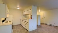 This single-floor space presents a practical and straightforward layout centered around an open kitchen area. The kitchen features white cabinetry with upper wall-mounted cabinets offering ample storage, complemented by a built-in sink set into a continuous countertop. A structural column with crown molding detail defines the kitchen's peninsula, creating a natural separation between the cooking area and the adjacent living space without fully closing off the floor plan.

The flooring throughout consists of neutral-toned square tiles, providing a durable and easy-to-maintain surface. Ceiling-mounted light fixtures illuminate both the kitchen and the adjoining hallway or entryway area, which is visible through the open layout and features a white paneled door with hardware. The walls are finished in a clean white throughout, contributing to a bright and cohesive aesthetic. A fire extinguisher is mounted near the kitchen workspace, adding a practical safety element. The overall design is functional and utilitarian, with a neutral color palette that creates a simple, unfussy atmosphere throughout the space.