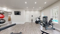 Located at 1173 North Hairston Road in Stone Mountain, GA, this fitness center offers a clean, modern workout environment designed to support a wide range of exercise routines. The space is housed on a single floor and provides a generous open layout, allowing ample room for equipment placement and freedom of movement throughout.

The interior features light gray luxury vinyl plank flooring, crisp white walls, and a neutral color palette that creates a bright, inviting atmosphere. Recessed lighting throughout the ceiling ensures consistent, well-distributed illumination across the entire workout area. Multiple large windows line the walls, flooding the space with natural light and offering views of the surrounding greenery outside.

The fitness center is outfitted with a comprehensive selection of equipment, including a treadmill, elliptical machine, rowing machine, leg press/extension machine, and a cable/functional trainer. A dedicated storage rack holds medicine balls, kettlebells, and resistance accessories, supporting functional and strength-based training. A wall-mounted flat-screen television is positioned for easy viewing during workouts.

Safety features include a clearly marked emergency exit sign and a fire extinguisher. Access to the exterior is available through a glass-panel door, and an interior door provides additional connectivity within the facility. The overall design prioritizes both functionality and comfort, making this a well-appointed fitness space.