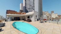 This rooftop pool space offers a compelling blend of outdoor recreation and indoor comfort, set against a striking urban skyline. The centerpiece is an oval swimming pool finished in vivid turquoise, surrounded by a spacious light-toned tile deck furnished with seating areas ideal for relaxing or socializing. The open-air terrace provides unobstructed views of the surrounding city high-rises, creating a dynamic backdrop throughout the day.

Adjacent to the pool deck, an interior lounge area extends the usable space with a clean, modern aesthetic. Light wood-look tile flooring runs throughout, complementing the crisp white walls and tray ceiling accented in soft blue-gray tones. Ceiling fans provide comfortable airflow, while large glass doors and windows connect the interior seamlessly to the outdoor terrace, allowing abundant natural light to fill the space.

The lounge is furnished with tufted leather sofas and armchairs arranged around a wall-mounted flat-screen television, creating a comfortable gathering space. A distinctive design detail is the blue mosaic tile accent surrounding the stairwell door, adding a splash of color and texture that echoes the hues of the pool. The overall ambiance is contemporary and polished, well-suited for both casual relaxation and social gatherings in an urban setting.