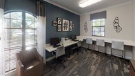 This single-floor business center combines functional workspaces with thoughtfully designed interior elements. The room is defined by a striking contrast between warm beige walls and a bold teal accent wall, both finished with elegant crown molding throughout. Patterned carpet in muted gray and blue tones anchors the space and adds visual texture underfoot.

A lounge area near the arched window features two upholstered swivel chairs positioned alongside a contemporary floor lamp, creating an informal seating nook bathed in natural light filtered through sheer curtains and horizontal blinds. The arched window serves as a notable architectural detail, lending a classic touch to the otherwise modern interior.

The workstation area lines the teal accent wall and accommodates multiple desktop computers and a printer along a continuous white surface-mounted desk. Ergonomic office chairs provide practical seating for focused tasks. A gallery-style arrangement of typographic framed prints decorates the wall above, adding a creative, editorial flair.

A separate alcove offers an additional white bench-height desk with paired seating and a sculptural black wall accent, ideal for quiet individual work. A white glass board panel provides an additional collaborative surface. Interior glass windows connect the space visually to an adjacent corridor, contributing to an open, connected feel.