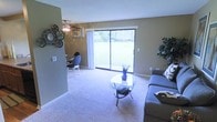 This single-floor residence offers a welcoming and functional layout centered around a bright, open-plan living and dining area. The living space is finished with plush wall-to-wall carpeting that adds warmth and comfort underfoot, while neutral-toned walls create a calm, versatile backdrop throughout. A dominant feature of the main living area is the large sliding glass door, which floods the interior with natural light and frames a picturesque view of the surrounding green landscape, seamlessly connecting the indoors with the outdoors. A ceiling fan with integrated lighting is mounted overhead, providing both air circulation and ambient illumination. An integrated wall-mounted air conditioning unit ensures year-round climate comfort. The dining area is situated adjacent to the living room in an open-concept arrangement, creating a cohesive and social atmosphere. Decorative wall accents, including a wall clock and circular metal art pieces, add character and a personalized touch to the space. The overall ambiance is relaxed and practical, with an emphasis on natural light, open sightlines, and comfortable living across the single level.