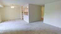 This single-floor residence is designed with a practical, open-concept layout that promotes a comfortable flow between living spaces. The main living area is finished with wall-to-wall neutral-toned carpet, lending a soft, understated aesthetic throughout. A ceiling fan is positioned overhead, providing both ventilation and a subtle decorative touch.

A partial wall divides the living area from the kitchen, creating a semi-open arrangement that maintains a sense of separation while preserving visual connectivity. The kitchen is equipped with white cabinetry, white appliances including a stove and refrigerator, and a sink positioned beneath overhead storage, offering a clean and functional workspace. The layout provides adequate counter space and storage for everyday use.

Adjacent to the main living area, a secondary room is visible through an opening, featuring hard-surface flooring that contrasts with the carpeted areas, adding variety to the interior finishes. The walls throughout are painted in a neutral off-white tone, contributing to a bright and airy atmosphere. The overall design is straightforward and move-in ready, suited to a range of living arrangements.