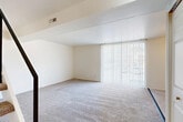 This two-floor residential space offers a clean, neutral aesthetic throughout, with soft neutral-toned carpet covering the living areas and bedrooms, and light tile flooring in the kitchen and bathroom. The main living area is generously proportioned, featuring a sliding glass door dressed with vertical blinds that allows abundant natural light to fill the room and provides views of the surrounding greenery. A structural beam detail adds architectural interest to the ceiling.

The kitchen is efficiently laid out with ample white cabinetry for storage, granite-style countertops, a white electric range, and a refrigerator. A ceiling fan with integrated lighting keeps the space comfortable. A large window beside the kitchen allows additional natural light to flow in.

The bathroom is compact and functional, featuring a bathtub and shower combination with white tile surround, a toilet, and tile flooring. The bedrooms are carpeted and bright, with clean white walls and wood-paneled ceilings that lend a distinct texture to the upper-floor rooms. An iron-railed staircase connects the two floors, adding a subtle design element. Overall, the space presents a bright, open, and functional layout well-suited for everyday living.