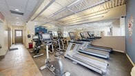 Located at 1414-1422 Som Center Rd in Mayfield Heights, Ohio, this fitness center offers a focused and functional workout environment within a single, efficiently organized space. The gym's architectural design features striking exposed steel roof trusses that lend an open, industrial character to the room, complemented by a mix of drop-ceiling sections and bright fluorescent lighting that keeps the space well-illuminated throughout.

The workout floor is equipped with a comprehensive selection of Matrix-brand cardio machines, including treadmills, ellipticals, upright bikes, and recumbent bikes, all arranged to maximize usability within the space. A large wall mirror along one side visually expands the room and allows exercisers to monitor their form. Natural light filters in through generous windows, creating an inviting and energizing atmosphere.

The flooring transitions between durable tile near the entry and carpeted exercise areas, providing practical surface variety. A bold motivational wall graphic reading "Walk In Strong, Walk Out Stronger" adds an encouraging, energetic ambiance. The space also includes a water fountain and restroom access, ensuring convenience for users. The overall design is clean, purposeful, and well-suited for a focused cardio-centric fitness experience.