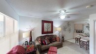 This single-floor space presents a welcoming living area characterized by a neutral color palette accented with rich, warm tones of red and brown throughout. The living room serves as the central gathering space, featuring a textured popcorn ceiling with a white ceiling fan that provides both light and airflow. The room accommodates comfortable seating, including a plush sofa adorned with decorative pillows in jewel tones, complementing the overall warm aesthetic.

Wall décor includes a bold red-framed artwork depicting a bare tree, adding a striking focal point, while ornamental metal wall art contributes additional visual interest. Decorative table lamps and floor lamps provide layered ambient lighting throughout the space. Red accent vases and floral arrangements reinforce the cohesive color scheme.

A light-toned carpet grounds the living area, adding softness underfoot, while a partial view into an adjacent room hints at a connected layout. The overall ambiance of this single-floor home is cozy and well-appointed, with thoughtful decorative touches that create a warm and lived-in atmosphere suitable for everyday comfort.