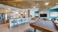 This well-appointed single-floor community space combines a kitchen and living room into one cohesive, open-plan layout designed for social gatherings and relaxation. The interior is defined by a striking slatted wood ceiling that adds warmth and texture throughout, complemented by polished stone tile flooring that flows seamlessly across the space.

The kitchen area features light wood cabinetry with gold hardware, open floating shelves, white quartz countertops, and a full suite of stainless steel appliances including a wall oven, dishwasher, and refrigerator. A large white bar counter with upholstered blue bar stools serves as a central gathering point and casual dining area.

The living zone is anchored by a teal accent wall paired with a large-format stone tile media wall housing a flat-screen TV. Plush terracotta lounge chairs and a cozy seating nook create an inviting atmosphere for unwinding. Pendant globe lights in brass fittings add a refined, modern touch overhead.

A regulation-size pool table sits prominently at the center of the room, making it an ideal entertainment hub. Large black-framed sliding glass doors line the exterior wall, flooding the space with natural light and providing direct access to an outdoor pool and patio area, seamlessly blending indoor and outdoor living.