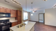 This single-floor residence showcases an open-concept layout that seamlessly connects the kitchen and living areas, creating a cohesive and functional living environment. The kitchen is appointed with rich, warm-toned wood cabinetry that extends to the ceiling, providing ample storage space. A large granite-topped island anchors the kitchen, featuring an undermount sink with a brushed gold faucet and pendant lighting suspended above, adding both practical task lighting and a decorative focal point. Track lighting along the kitchen ceiling ensures the workspace is well-illuminated. The transition into the living area is smooth and natural, where a ceiling fan with a light fixture maintains airflow and comfort throughout the open space. Dark hardwood-style flooring runs consistently through both the kitchen and living areas, grounding the warm, neutral color palette of the walls. A window in the living area allows natural light to filter into the space. A doorway visible from the main living area leads to an adjacent room with carpeted flooring, suggesting a private bedroom or secondary space. The overall ambiance is clean, modern, and welcoming.