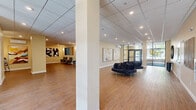 Located at 268 Market St in South Burlington, Vermont, this ground-floor commercial space is part of a modern, multi-story mixed-use building situated along a vibrant, tree-lined streetscape. The building's exterior showcases a striking combination of brick, wood panel, and metal cladding, with large black-framed windows that lend a contemporary industrial character. Accessible from both the street-facing sidewalk and a rear parking area, the space features a welcoming entry with bold graphic signage on the glass facade.

Inside, the generously sized interior is organized across an open, flowing layout finished with warm wood-look flooring, recessed lighting, and a suspended tile ceiling. The main lobby area is appointed with curved, plush seating arrangements and an impressive collection of curated wall art—including large abstract paintings and a striking three-dimensional textured wall piece—creating an inviting, gallery-like atmosphere. A secondary lounge area provides an intimate setting with high-back upholstered chairs, a side table, and additional artwork displays.

Steel-framed glass partition walls define interior zones while maintaining visual openness and allowing natural light to filter throughout. The overall ambiance is polished yet welcoming, making this space well-suited for a professional, community-facing, or hospitality-oriented use.