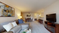 Located at 5032 S Falcon Glen Blvd in Greenfield, Wisconsin, this single-floor residence offers a thoughtfully designed open-concept layout that seamlessly connects the living, dining, and kitchen areas. The living room features plush wall-to-wall carpeting and is furnished with a comfortable sectional sofa and accent chair, accented by warm ambient lighting that creates an inviting atmosphere. Adjacent to the living area, the dining space accommodates a full six-person table beneath a pendant light fixture, while a raised breakfast bar with barstool seating provides an additional casual dining option. The kitchen is appointed with rich cherry wood cabinetry, black appliances including a range, microwave, and refrigerator, and durable hardwood-style flooring that adds warmth and contrast. Two well-proportioned bedrooms are finished with soft carpeting, ample closet space, and natural light from well-placed windows. Each bedroom is served by its own full bathroom, both featuring white cabinetry vanities, tile flooring, and clean, neutral finishes. Track lighting in the common areas and consistent warm tones throughout the home contribute to a cohesive, comfortable ambiance suited for everyday living.