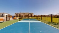 This outdoor recreational facility features two dedicated pickleball courts situated side by side, offering ample space for both casual play and competitive matches. The courts are finished in a striking two-tone blue surface with clearly marked white boundary lines and light blue non-volley zones, set against a green perimeter surround that enhances visibility and playability. Each court is equipped with a regulation net, and the entire facility is enclosed within sturdy black metal fencing that defines the playing area while maintaining an open, airy feel. Gated entry points provide easy access from multiple sides. Wooden benches are positioned along the interior perimeter of the courts, offering comfortable seating for players waiting their turn or spectators observing the action. Beyond the fencing, well-maintained green lawns extend outward, with views of a multi-story residential building complex and neighboring rooftops visible in the background. The surrounding landscape includes young trees and open sky, contributing to a bright, sun-drenched atmosphere. The facility is thoughtfully designed to accommodate multiple players simultaneously, making it an ideal amenity for an active residential community. The clean, well-maintained surfaces and organized layout reflect a commitment to quality outdoor recreation.