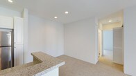 This single-floor space offers a clean, contemporary design characterized by crisp white walls, recessed ceiling lighting, and plush neutral-toned carpeting that creates a warm and inviting atmosphere throughout. The open layout seamlessly connects the kitchen and living areas, promoting a sense of spaciousness and ease of movement. A standout feature is the granite-topped breakfast bar, which serves as both a functional surface and a natural dividing element between the kitchen and the adjoining living space. The granite countertop showcases subtle veining in soft earth tones, adding a touch of elegance to the otherwise minimalist aesthetic. Recessed lighting is strategically positioned in the ceiling to provide even, ambient illumination across the main living area. A hallway visible from the main space leads to additional rooms, suggesting a well-organized and practical floor plan. The overall design philosophy of this space leans toward simplicity and functionality, with a neutral color palette that provides a versatile backdrop adaptable to a variety of interior styles. The combination of clean lines, quality finishes, and thoughtful layout makes this a highly functional and visually cohesive space.