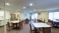 This single-floor communal space is thoughtfully designed to accommodate group dining or social gatherings in a comfortable and accessible setting. The room features multiple rectangular white-top tables, each surrounded by sturdy brown upholstered chairs, arranged throughout the space to encourage both intimate conversations and larger group interactions. The flooring consists of a patterned carpet that adds warmth and texture to the overall environment. Recessed ceiling lights are evenly distributed across the room, providing consistent, well-distributed illumination throughout the space. A large window along the far wall allows natural light to fill the room, offering views of the greenery outside and creating a bright, pleasant ambiance. A wall-mounted air conditioning unit ensures year-round comfort for occupants. The walls are painted in a soft, neutral tone and adorned with framed artwork, contributing to a calm and welcoming atmosphere. A mirrored wall feature near the entrance enhances the sense of openness and adds a decorative touch. The overall design prioritizes functionality and ease of movement, making this a practical and inviting space suited for daily communal use.