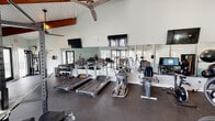 Located at 1800 William Kennerty Dr in Charleston, South Carolina, this well-equipped fitness center is housed in a single-story facility distinguished by its striking vaulted ceilings with exposed wood beams and white tongue-and-groove paneling. The architecture creates an open, airy atmosphere that is further enhanced by large glass doors and windows that allow natural light to pour in from multiple sides, offering views of the surrounding outdoor greenery.

The gym floor is covered with durable rubber flooring, providing a safe and functional surface throughout. A full wall of mirrors expands the sense of space and allows users to monitor their form during workouts. Multiple wall-mounted flat-screen televisions are positioned for visibility from across the room, and large ceiling fans maintain airflow and comfort.

The cardio section is outfitted with a row of Life Fitness treadmills, elliptical trainers, and stationary and recumbent bikes. The strength training area features a multi-function cable machine with suspension workout attachments, a power rack with weight plates, a comprehensive dumbbell rack ranging across multiple weight increments, and a selection of medicine and stability balls. Workout reference charts are posted on the walls for added guidance. The space is thoughtfully organized to accommodate a variety of training styles in a comfortable, well-lit environment.