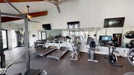 Located at 1800 William Kennerty Dr in Charleston, South Carolina, this well-equipped fitness center is housed in a single-story facility distinguished by its striking vaulted ceilings with exposed wood beams and white tongue-and-groove paneling. The architecture creates an open, airy atmosphere that is further enhanced by large glass doors and windows that allow natural light to pour in from multiple sides, offering views of the surrounding outdoor greenery.

The gym floor is covered with durable rubber flooring, providing a safe and functional surface throughout. A full wall of mirrors expands the sense of space and allows users to monitor their form during workouts. Multiple wall-mounted flat-screen televisions are positioned for visibility from across the room, and large ceiling fans maintain airflow and comfort.

The cardio section is outfitted with a row of Life Fitness treadmills, elliptical trainers, and stationary and recumbent bikes. The strength training area features a multi-function cable machine with suspension workout attachments, a power rack with weight plates, a comprehensive dumbbell rack ranging across multiple weight increments, and a selection of medicine and stability balls. Workout reference charts are posted on the walls for added guidance. The space is thoughtfully organized to accommodate a variety of training styles in a comfortable, well-lit environment.