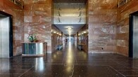 Located at 227 Church Street in New Haven, CT, this grand lobby is a masterclass in Art Deco design. The elongated, corridor-style space is clad floor-to-ceiling in richly veined rose and amber marble, creating a warm, distinguished atmosphere. Polished dark terrazzo floors feature intricate inlaid white line work depicting a geographic map, adding a unique and historically significant artistic element underfoot.

The lobby is flanked on both sides by a series of stainless steel elevator doors, each adorned with decorative motifs including stylized bird figures, geometric patterns, and circular port-style detailing. Flush dome ceiling fixtures cast a soft, even glow along the full length of the corridor. At the far entry vestibule, two elegant revolving doors with bronze and glass construction invite guests in, set within a mirrored, glass-paneled foyer that reflects abundant natural light.

At the opposite end, a decorative metal panel featuring seabirds in flight serves as a striking focal point above the corridor entrance. Coffered ceiling sections with gilded, geometric Art Deco friezes frame the elevator alcoves, adding to the architectural richness. A modest reception counter and floral arrangement provide a welcoming human touch within this otherwise monumental space.
