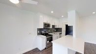 This single-floor space showcases a thoughtfully designed kitchen that serves as the heart of the home. The kitchen features crisp white shaker-style cabinetry that extends to the ceiling, providing generous storage and a clean, timeless aesthetic. A dark grey subway tile backsplash offers a striking contrast against the white cabinetry and light quartz countertops, adding depth and visual interest to the space. Stainless steel appliances, including a range with oven, over-the-range microwave, dishwasher, and a full-size refrigerator, lend a modern, professional feel to the kitchen. A peninsula with a seamless white countertop extends the workspace and creates a natural division between the kitchen and the adjacent living area, offering additional seating or prep space. Recessed lighting illuminates the space efficiently, complementing the natural light that filters in from above. The warm-toned hardwood-style flooring runs throughout, grounding the bright, neutral palette and adding warmth to the overall ambiance. The space is finished with clean white walls and crisp baseboards, creating a fresh, move-in-ready environment that balances modern style with functional design.