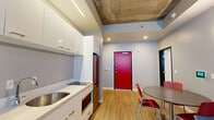 This thoughtfully designed two-bedroom, two-bathroom unit offers a compact yet functional layout across a single floor. The open-concept kitchen and living area serves as the heart of the space, characterized by an eye-catching exposed concrete ceiling that lends an industrial aesthetic. The kitchen features white cabinetry, a stainless steel sink, induction cooktop, and stainless steel refrigerator, all finished with light quartz countertops. A bold red entry door adds a striking focal point to the common area, which also accommodates a dining table and seating area with a window providing natural light.

The living area extends toward a window-facing seating arrangement, complete with a wall-mounted television, creating a comfortable gathering space. Light wood-tone flooring runs consistently throughout the unit, contributing to a clean, cohesive look.

Both bedrooms are modestly scaled, each featuring a window with blinds for natural light and privacy, along with built-in storage solutions including cabinetry and drawers. The two bathrooms are efficiently designed with white vanities, under-sink cabinetry, wall-mounted medicine cabinets, and sliding barn-style doors. A connecting hallway links the bedrooms and bathrooms, ensuring a practical flow throughout the unit.