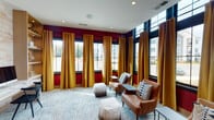 Located on Bonnar Bridge Parkway in Burlington, North Carolina, this single-floor amenity space combines vibrant design with practical functionality. The lounge area makes an immediate impression with its bold crimson accent walls, floor-to-ceiling black-framed windows, and tall golden-yellow drapery panels that frame sweeping views of the surrounding community. Abundant natural light fills the room, highlighting the plush patterned area rug, cognac leather armchairs with geometric throw pillows, and faux fur ottomans arranged for casual conversation. The elevated ceiling height enhances the open, airy feel of the space.

Adjacent to the lounge, a well-appointed business center offers a long, built-in white desk topped with multiple desktop computers and a wall-mounted flat-screen television. A textured, light-toned wood-panel accent wall provides a warm backdrop, while built-in shelving units with integrated lighting offer both display space and functional storage. Black modern task chairs complete the workspace. The two areas connect seamlessly through a glass-panel partition, maintaining visual openness while defining distinct zones. The overall design balances bold color and contemporary furnishings to create a welcoming, polished environment.