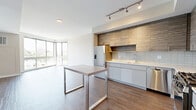 Located at 900 Pacific St in Stamford, Connecticut, this modern one-bedroom, one-bathroom residence offers a well-designed layout across a single floor. The open-plan living and kitchen area is bathed in natural light, courtesy of striking floor-to-ceiling corner windows that frame views of the surrounding Stamford cityscape and greenery, creating a bright and airy atmosphere throughout the day.

The kitchen is thoughtfully appointed with two-tone cabinetry — combining warm wood-grain uppers with sleek white lower cabinets — complemented by a white subway tile backsplash and a full suite of stainless steel appliances, including a gas range, dishwasher, microwave, and refrigerator. A peninsula-style table with metal legs provides additional workspace and casual dining.

The bedroom is a well-proportioned retreat featuring its own large window with outdoor views and warm wood-tone flooring that carries throughout the unit. A dedicated closet offers practical storage.

The bathroom is finished in a clean, contemporary style with wide-format wood-look flooring, a floating vanity with an illuminated mirror, and a glass-enclosed shower. Neutral tones and recessed lighting lend the space a polished, spa-like quality.

Warm wood-look flooring, neutral walls, and clean-lined architectural details unify the space, giving it a cohesive modern aesthetic.