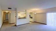 This single-floor residence features an open-concept design that seamlessly connects the living, dining, and kitchen areas into one cohesive space. The living area is finished with neutral wall-to-wall carpeting, creating a warm and quiet underfoot feel, while plain white walls provide a clean, versatile backdrop throughout.

The kitchen is positioned behind a half-wall pass-through counter, offering both separation and connectivity to the main living space. White cabinetry lines the upper and lower sections of the kitchen, complemented by a dark countertop with an integrated sink. A refrigerator is visible to the side, and built-in appliances are neatly tucked beneath the upper cabinets, making the kitchen both functional and well-organized.

Adjacent to the kitchen pass-through, a dedicated dining area is defined by a ceiling-mounted pendant light fixture, which adds a subtle focal point to the space. A mirrored accent panel on the dining area wall enhances the sense of depth and reflects light throughout the room.

Overall, the layout is practical and straightforward, with a neutral color palette and standard finishes that lend the space a clean, move-in-ready quality.