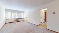 This single-floor unit is laid out with a functional and straightforward design. The living area is generously proportioned, featuring neutral wall-to-wall carpeting, crisp white walls, and a large multi-pane window fitted with horizontal blinds that allows ample natural light to fill the room. A wall-unit air conditioner and baseboard heating system provide year-round climate control. The open layout connects seamlessly to a secondary sitting or dining area through a wide doorway.

The galley-style kitchen runs parallel walls of white cabinetry with round knob hardware and laminate countertops. A stainless steel double sink, a white electric range, and a full-size refrigerator are included. Light tile flooring and under-cabinet lighting add practicality to the space.

The unit includes two carpeted bedrooms. The primary bedroom benefits from dual windows with blinds and a ceiling fan, while the secondary bedroom features a single window with a baseboard heater beneath it. Both rooms offer a calm, neutral palette.

The bathroom retains a classic retro aesthetic, showcasing pink ceramic wall tiles, a mosaic tile floor, and a matching tub and sink. A built-in storage cabinet and entry closet with bifold doors round out the unit's storage offerings.