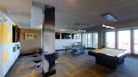 This single-floor recreational space is a well-appointed game room designed for social entertainment and leisure. The room's most distinctive architectural feature is a bold structural column wrapped in a baseball graphic, serving as a striking visual centerpiece that sets the sports-inspired tone throughout the space. The layout accommodates multiple activities simultaneously, with a full-size ping pong table positioned centrally and a pool table situated nearby, offering ample room for play without crowding. A lounge area with circular bar-height stools provides casual seating for spectators or relaxed conversation. Mounted flat-screen televisions are positioned for clear sightlines from multiple vantage points, enhancing the entertainment experience. Large floor-to-ceiling windows along one wall flood the space with natural light and offer expansive exterior views, creating an open and energetic ambiance. The flooring features a multi-toned carpet tile in a geometric pattern that adds visual texture and defines the space. Overhead, a combination of recessed lighting and a modern pendant fixture provide layered illumination. The overall design balances an athletic aesthetic with modern amenity finishes, making it a versatile and inviting destination for recreation and social gatherings.